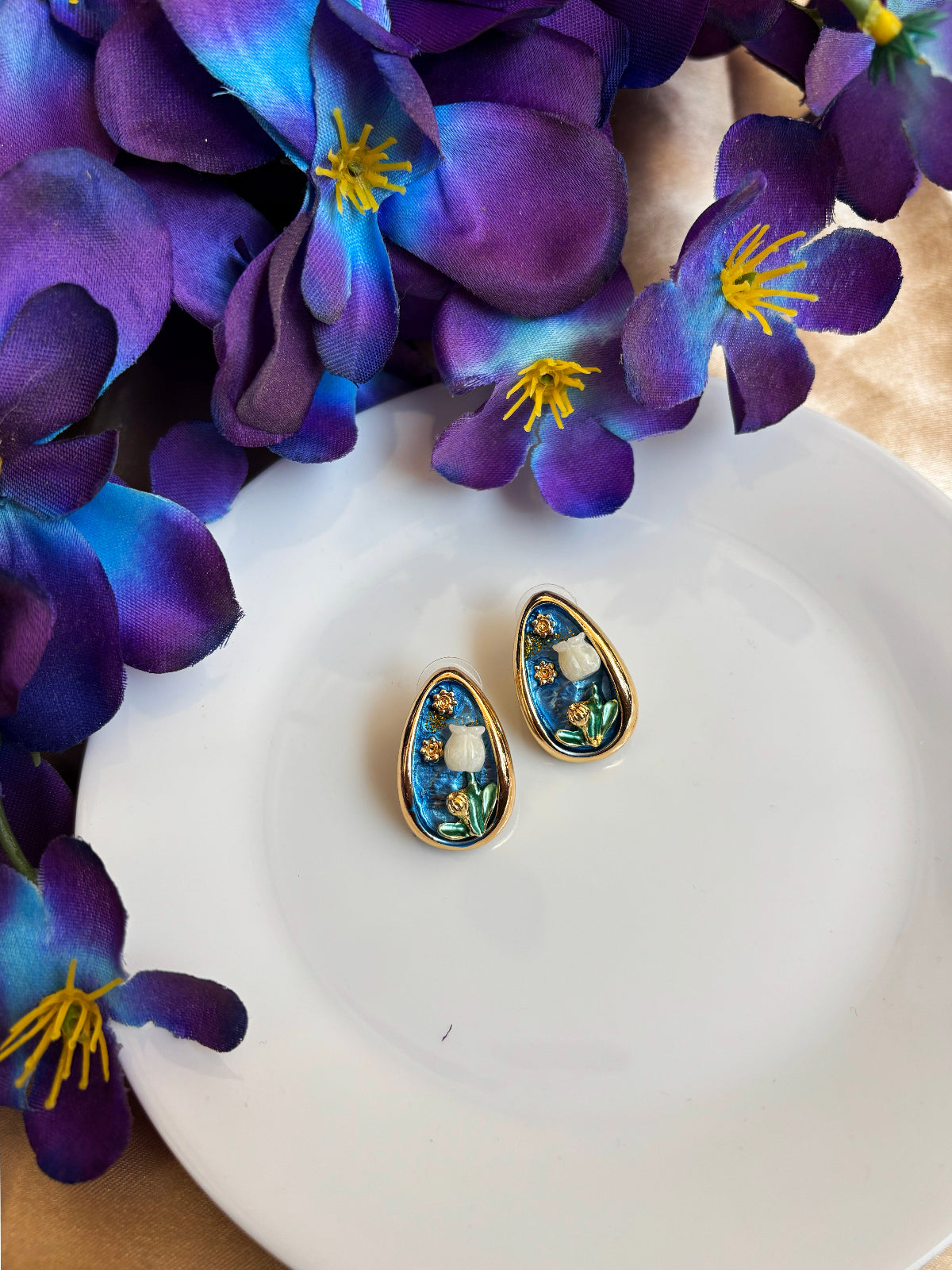 Teardrop-shaped earrings with floral design on a white plate surrounded by purple flowers.
